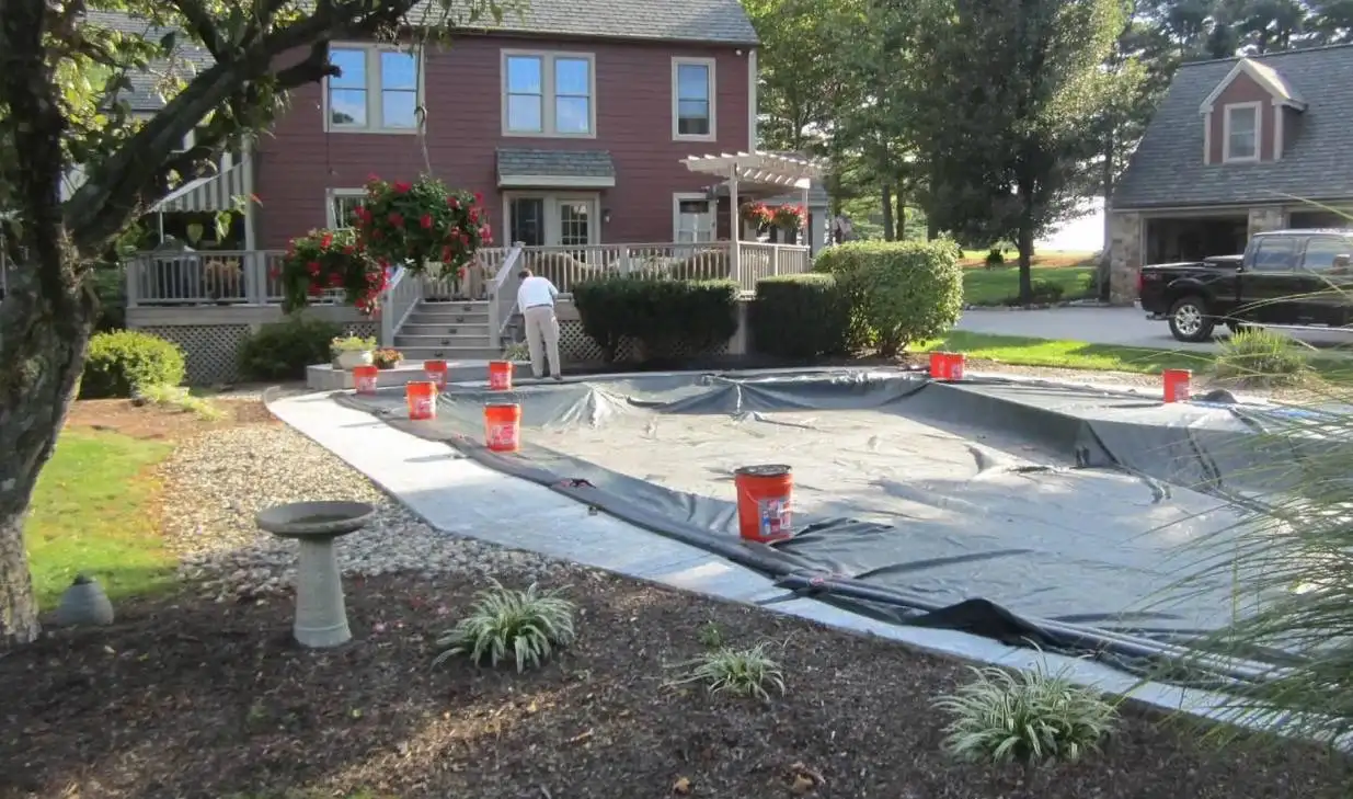 A covered pool with buckets on the edges sits in a backyard where a man adjusts the cover. The yard features a red house with a flower-adorned porch, a small garage, and is surrounded by trees and shrubs. This serene PA setting highlights expert pool construction in Delaware County, with a birdbath in the foreground.